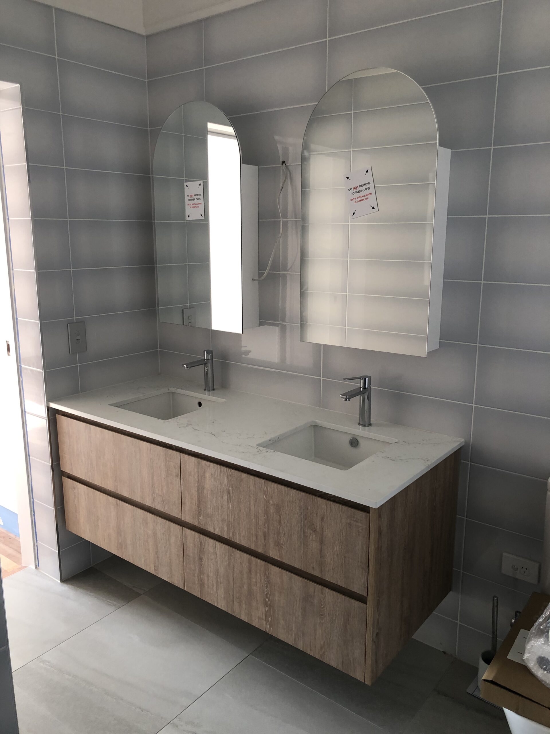 Modern bathroom with a floating wooden vanity, dual sinks, marble countertop, arched mirrored cabinets, and light grey tiled walls in Port Douglas.